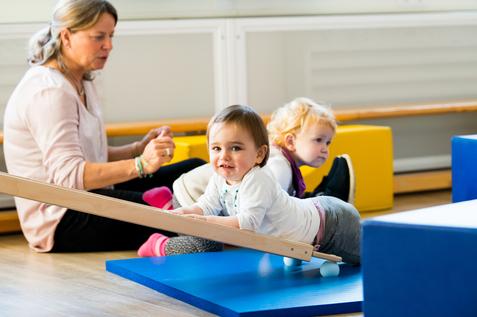Zwei Kleinkinder spielen auf einer blauen Matte, während eine betreuende Person im Hintergrund sitzt.